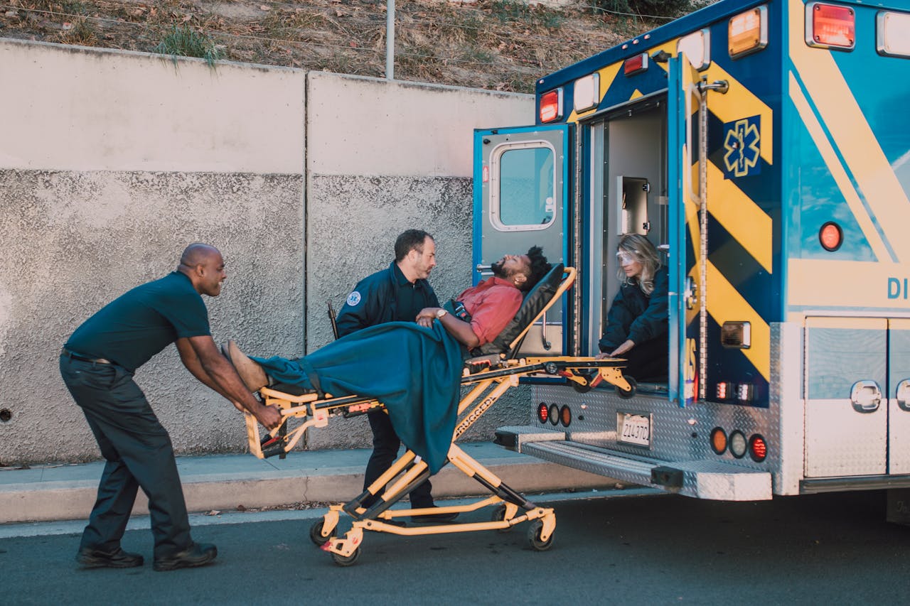 First responders assist an injured man onto a stretcher and into an ambulance during a daytime emergency call.