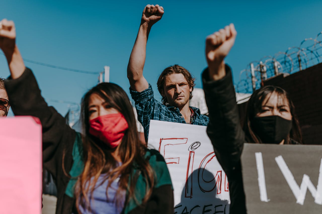 A diverse group of protesters demonstrate with raised fists and placards outdoors.